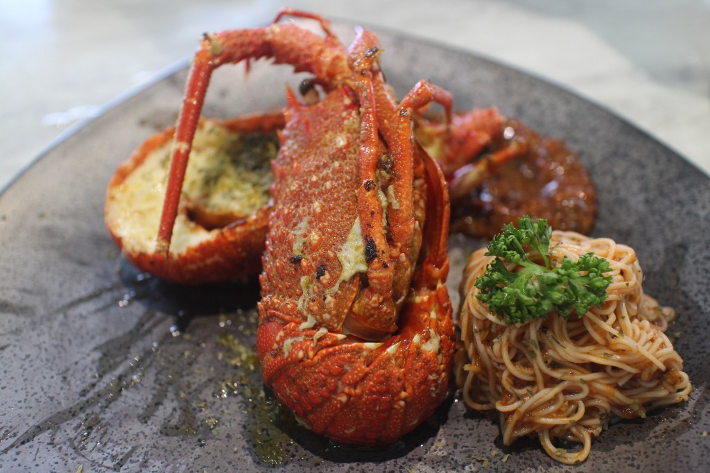 香煎龍蝦搭配番茄紅醬與澎湖麵線,淋上新鮮調味,擺放於黑色盤子上,旁邊有裝飾的香菜。