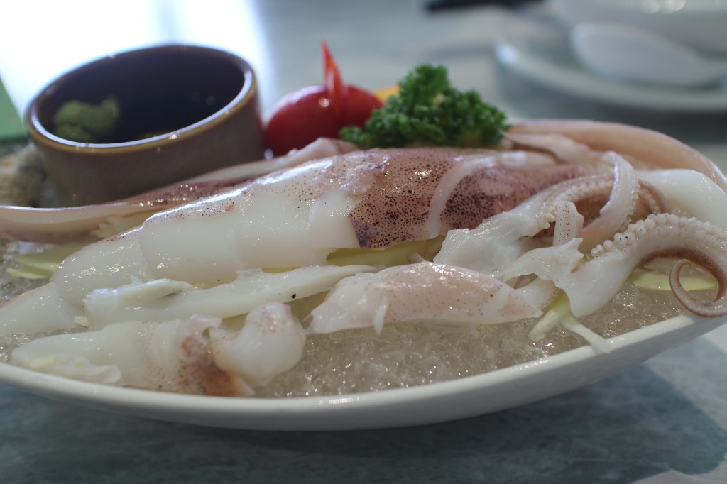 一盤新鮮的生食海鮮拼盤,包含各種切片的魷魚,搭配綠色芥末醬和醬油,旁邊有一片小番茄和一小塊香菜裝飾。